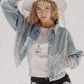 a young woman wearing a denim jacket with rhinestone fringe, black denim shorts, and a white western hat