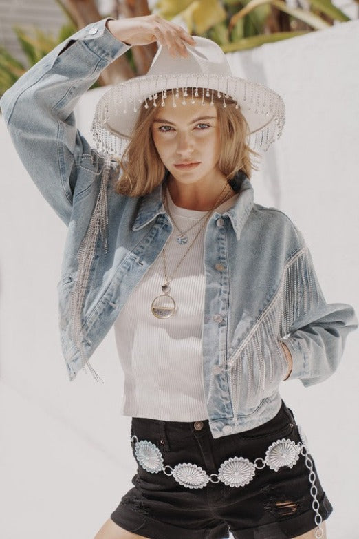 a young woman wearing a denim jacket with rhinestone fringe, black denim shorts, and a white western hat